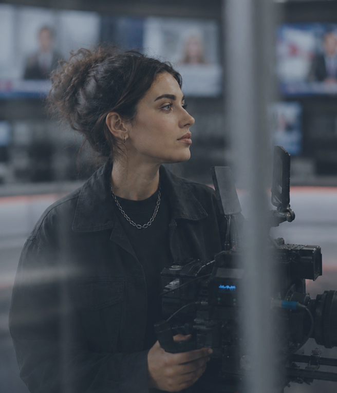 Woman with a focused expression holds a camera in a busy newsroom. Screens are blurred in the background.