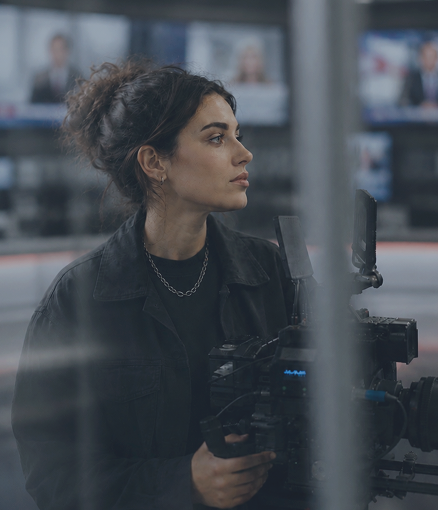 Woman with a focused expression holds a camera in a busy newsroom. Screens are blurred in the background.